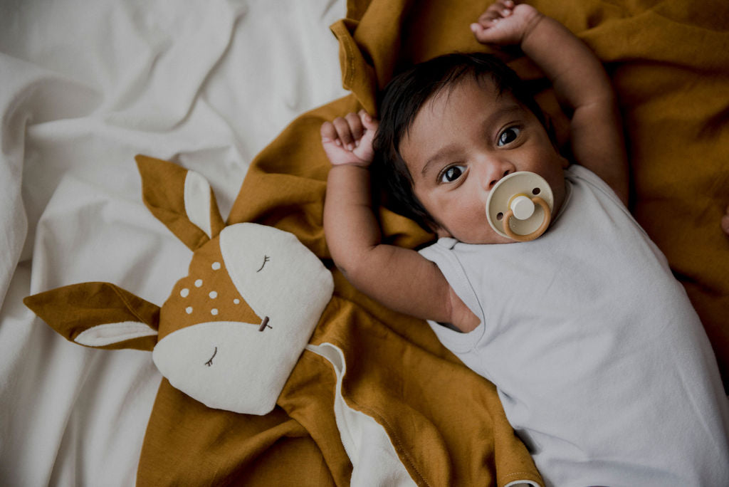 A content baby with a pacifier laying on a linen blanket with a Little Koko Deer comforter